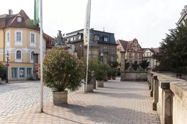 Pflasterplatz mit Blumenk&uuml;beln und Fahnenstangen, mehrst&ouml;ckige historische Geb&auml;ude im Hintergrund, Steinbalustrade rechts, bew&ouml;lkter Himmel, keine Personen