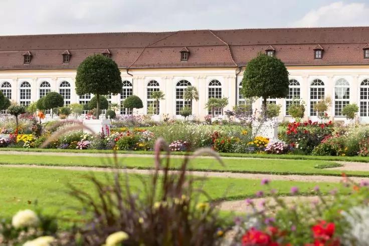 Vordergrund unscharfe Pflanzen, mittig Rasenfl&auml;chen und Kieswege, bunte Blumenbeete, im Hintergrund langes wei&szlig;es Geb&auml;ude mit vielen Rundbogenfenstern und rotem Ziegeldach, zwei kugelf&ouml;rmig geschnittene B&auml;ume