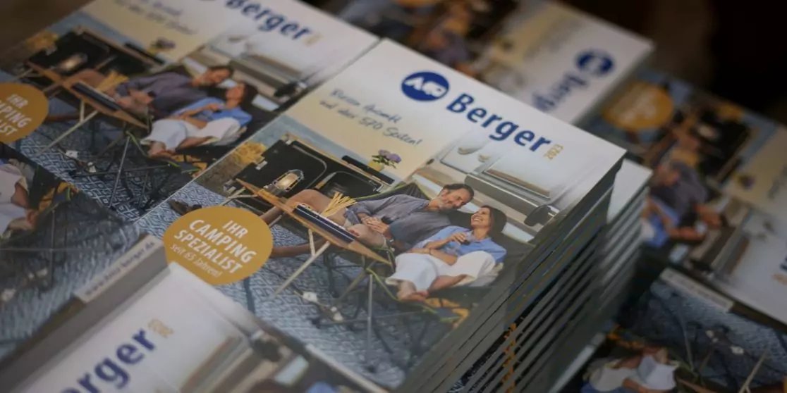 Gestapelte Brosch&uuml;ren mit blauem "Berger"-Logo; auf dem Cover zwei Personen, die an einem Holztisch sitzen, Getr&auml;nke auf dem Tisch und ein Fahrrad im Hintergrund; runder gelber Aufkleber auf dem Cover