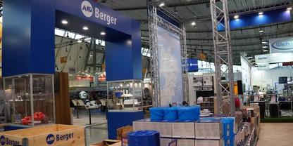 Blaues Schild mit Schrift 'AB Berger' &uuml;ber einem Bereich, Stapel blauer Kunststoffbeh&auml;lter und Kartons auf Paletten, Metalltr&auml;ger und Deckenbeleuchtung in einer Halle
