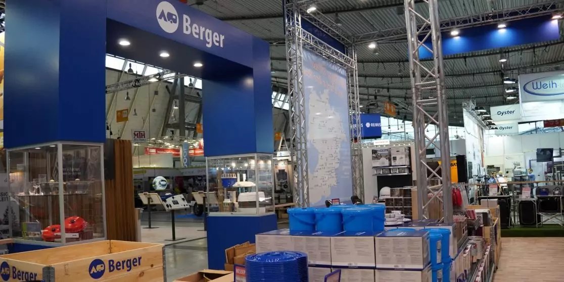 Blaues Schild mit Schrift 'AB Berger' &uuml;ber einem Bereich, Stapel blauer Kunststoffbeh&auml;lter und Kartons auf Paletten, Metalltr&auml;ger und Deckenbeleuchtung in einer Halle