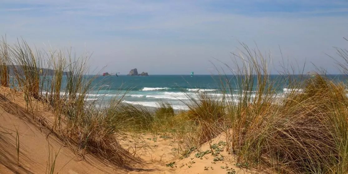 Sandd&uuml;nen mit Strandhafer und einem Pfad zum Meer, kleine Wellen, Felsformationen in der Ferne, blauer Himmel
