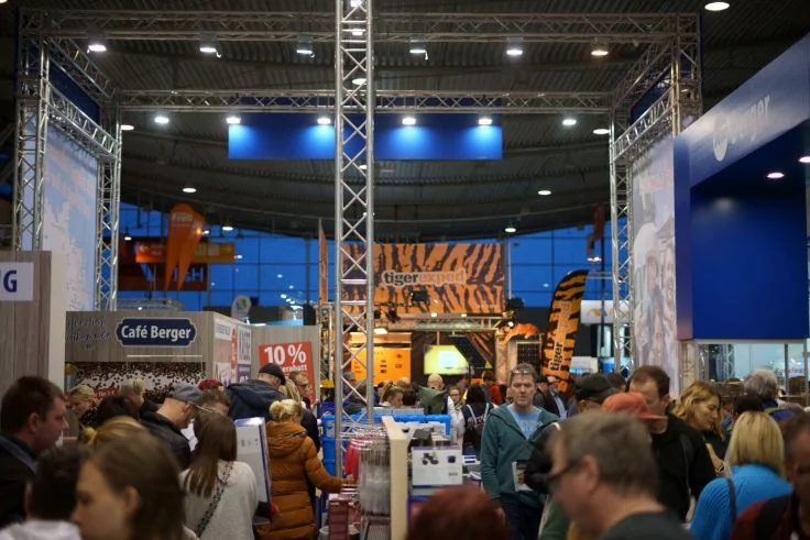 Innenraum mit Messeständen und Metallträgern, viele Menschen in den Gängen, links Schild 'Café Berger' und rotes Schild '10%', hinten großes Banner mit Tigerstreifen