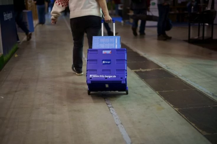 Person zieht eine blaue Kunststoffbox auf Rollen über den Hallenboden mit weißer Linie; weitere Personen unscharf im Hintergrund