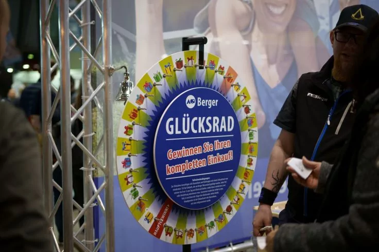 Blick auf ein aufgestelltes Glücksrad mit blauer Mittelplatte und Schrift 'Glücksrad' sowie 'Gewinnen Sie Ihren kompletten Einkauf!', farbige Segmente mit Geschenk-Icons. Rechts und links stehen zwei Personen, im Hintergrund ein Metallträger und ein