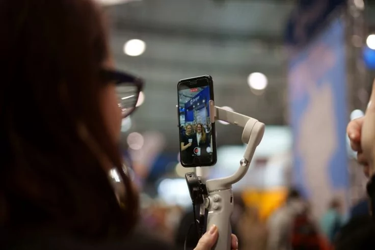 Person hält ein Smartphone auf einem Gimbal-Stabilisator; auf dem Display ist ein Bild von zwei Personen sichtbar; im Hintergrund unscharfe Menschen, Messestand und Deckenlichter