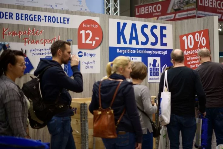 Mehrere Personen stehen in einer Schlange vor einem Messestand mit großem Schild 'KASSE'; Plakate zeigen '12,-' und '10%'
