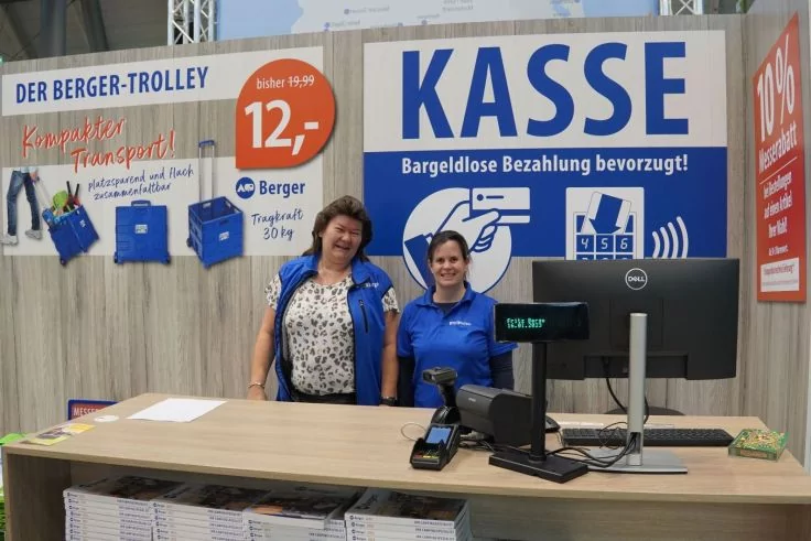Zwei Personen in blauer Kleidung stehen hinter einem Holzkassen-Tresen mit Kassenmonitor und Kartenleser; darüber großes Schild 'KASSE'; sichtbare Preisschilder '12,-' und '10%'