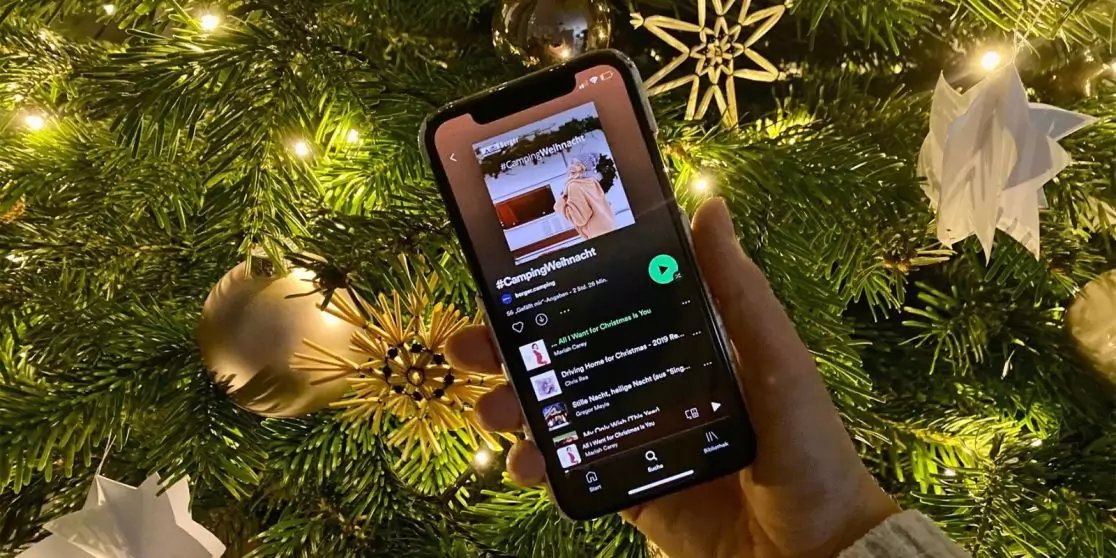 Hand h&auml;lt Smartphone mit Musikplayer-Bildschirm (Albumcover, Titelliste, Wiedergabeknopf); im Hintergrund geschm&uuml;ckter Tannenbaum mit Lichtern und Sternornamenten