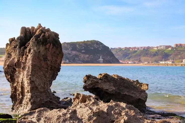 Felsformationen im Vordergrund, ruhiges Meer, weiter Klippen mit kleinem wei&szlig;em Leuchtturm und H&auml;usern unter blauem Himmel