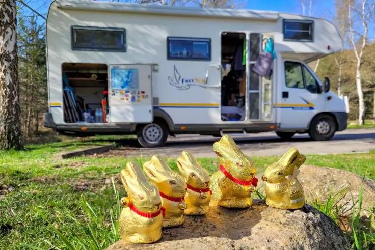 Vier goldene Hasen in Folienverpackung mit roten B&auml;ndern auf einem Stein im Vordergrund; im Hintergrund ein geparktes Wohnmobil mit offener T&uuml;r und ge&ouml;ffnetem Seitenfach, B&auml;ume und Gras