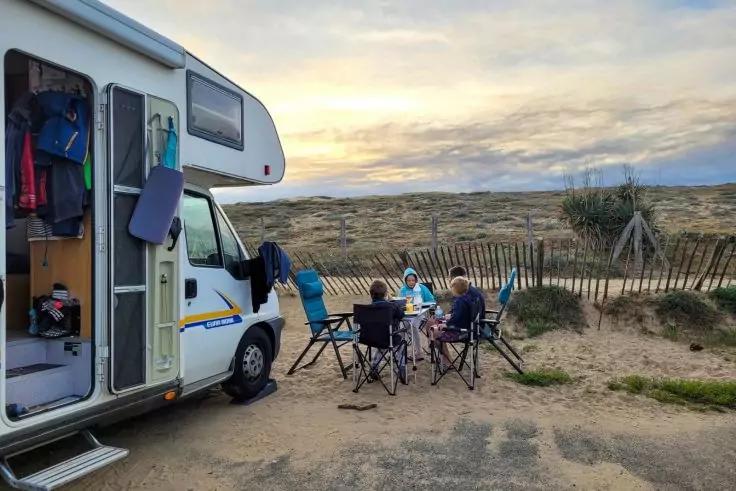 Wohnmobil mit ge&ouml;ffneter T&uuml;r und h&auml;ngender Kleidung; vier Personen sitzen an einem Klapptisch mit St&uuml;hlen vor D&uuml;nen und Holzzaun bei Sonnenuntergang