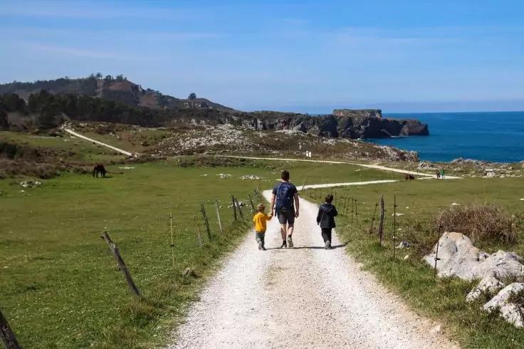 Drei Personen gehen mit dem R&uuml;cken zur Kamera auf einem Kiesweg zur K&uuml;ste, umgeben von gr&uuml;nen Wiesen, Felsen und blauem Himmel