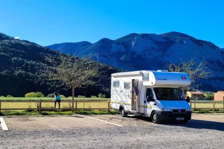 Wei&szlig;es Wohnmobil auf einem Parkplatz vor gr&uuml;nen Wiesen und Bergen; links eine Person neben einem Holzzaun, klarer blauer Himmel