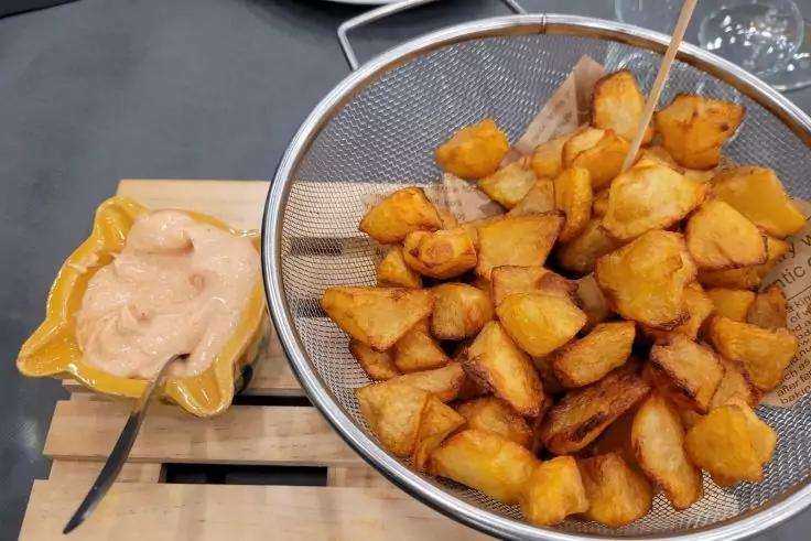 Metallsieb mit goldbraunen, gew&uuml;rfelten Kartoffelst&uuml;cken und Holzspie&szlig;; daneben gelbe Schale mit hellrosa Sauce und L&ouml;ffel auf einer Holzunterlage, dunkle Tischfl&auml;che im Hintergrund