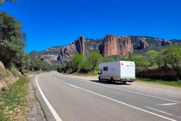 Wei&szlig;es Wohnmobil auf einer Landstra&szlig;e, rechts B&auml;ume und Str&auml;ucher, im Hintergrund rote Felsklippen unter blauem Himmel