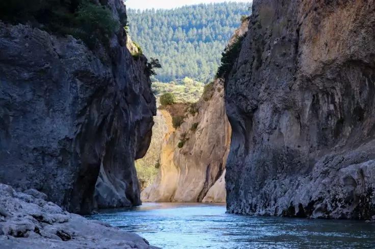 Schmaler Fluss zwischen hohen Felsw&auml;nden, steiniger Uferbereich im Vordergrund, bewaldeter Bergr&uuml;cken im Hintergrund