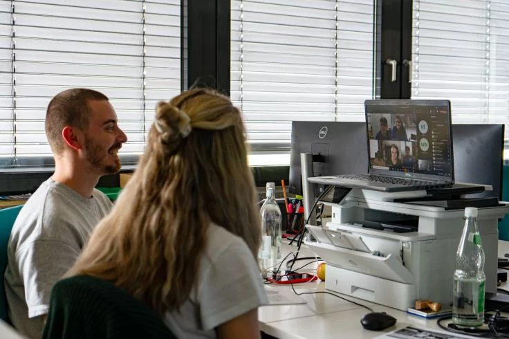 Zwei Personen an einem Schreibtisch, Monitor zeigt eine Videokonferenz, Drucker und Getr&auml;nkeflaschen auf dem Tisch, Jalousien im Hintergrund