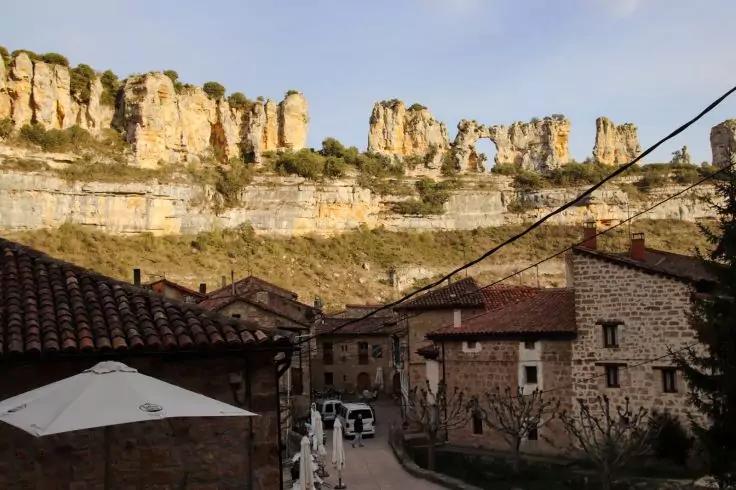 Schmale Gasse mit steinernen H&auml;usern, Au&szlig;ensitzbereich mit wei&szlig;en Sonnenschirmen und geparktem Auto, im Hintergrund hohe Felsklippen mit einem Felsbogen unter blauem Himmel