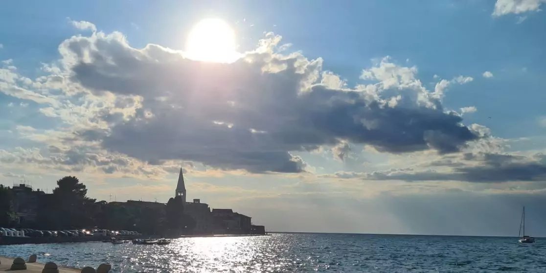 K&uuml;stenorts-Silhouette mit Kirchturm links, Sonne hinter Wolken &uuml;ber dem Meer, Lichtspiegelungen auf dem Wasser und ein kleines Segelboot rechts