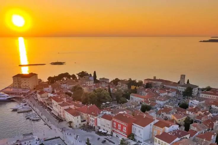 Sonnenuntergang &uuml;ber ruhiger See, Sonne nahe dem Horizont mit Lichtspiegelung, K&uuml;stenstadt mit dicht stehenden H&auml;usern und roten D&auml;chern, Uferpromenade und vereinzelten Booten