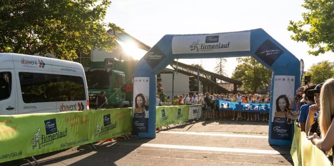 Aufblasbarer blau-wei&szlig;er Torbogen &uuml;ber einer Stra&szlig;e, Menschen hinter gr&uuml;nen Absperrbannern, wei&szlig;er Kleinbus links, B&auml;ume und Sonnenschein