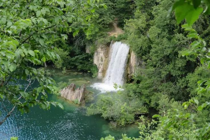 Wasserfall flie&szlig;t von einer Felswand in einen gr&uuml;n-t&uuml;rkisen See, umgeben von dichtem Laub; im Vordergrund Bl&auml;tter rahmen die Szene