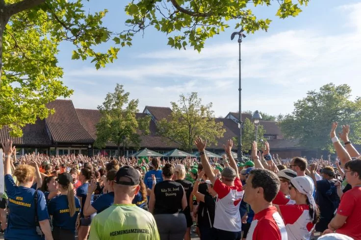 Menschenmenge im Freien, viele Personen von hinten mit erhobenen Armen, B&auml;ume, Geb&auml;ude mit Satteld&auml;chern, Laterne und blauer Himmel
