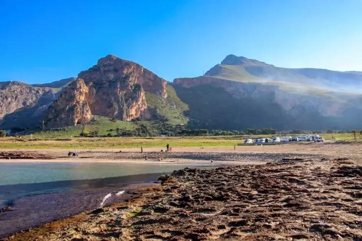 Sandiger Strand mit seichtem Wasser im Vordergrund, dahinter Berge und blauer Himmel, einzelne Personen, kleine Geb&auml;ude und ein Vogel am Ufer