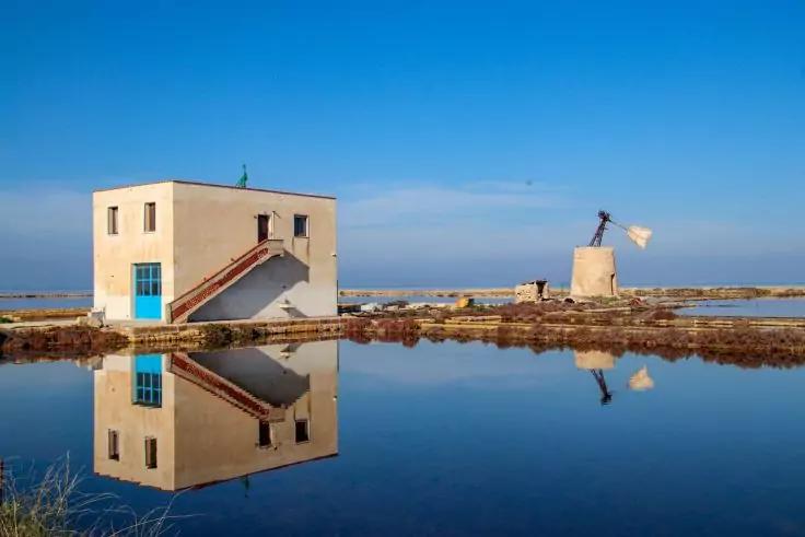 Rechteckiges helles Geb&auml;ude mit Au&szlig;entreppe und blauem Fenster/T&uuml;r neben einem kleinen runden M&uuml;hlenturm; beide spiegeln sich in ruhigem Wasser, flaches Land und klarer blauer Himmel
