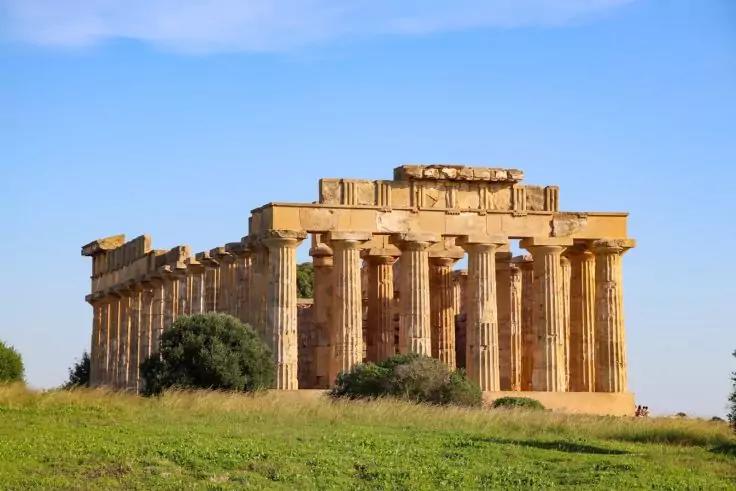 Rechteckiges steinernes Bauwerk mit hohen, zylindrischen S&auml;ulen in Reihen und teilweise fehlendem Dach auf einer gr&uuml;nen Wiese unter blauem Himmel