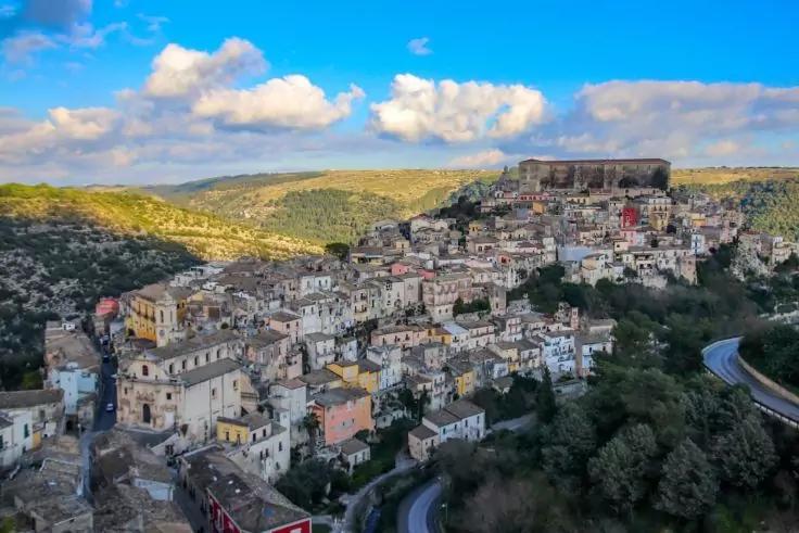 Siedlung auf einem H&uuml;gel mit dicht aneinander gebauten hellen H&auml;usern, gro&szlig;em Geb&auml;ude auf der Anh&ouml;he, kurviger Stra&szlig;e, B&auml;umen und blauem Himmel mit Wolken