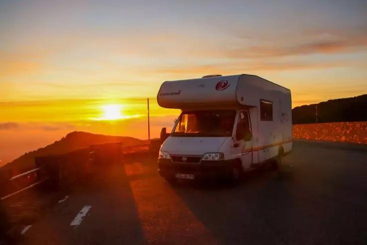 Wei&szlig;es Wohnmobil steht auf einer Stra&szlig;e neben einer Leitplanke, im Hintergrund Sonnenuntergang mit orange-gelbem Himmel und H&uuml;geln