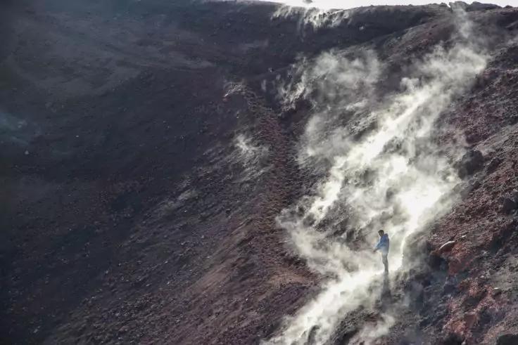 Einzelne Person in blauer Jacke auf einem dunklen, felsigen Hang; wei&szlig;e Dampfwolken steigen vom Boden auf