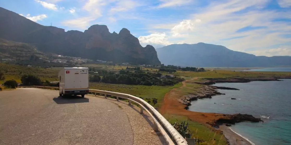 Wei&szlig;es Wohnmobil auf kurviger K&uuml;stenstra&szlig;e mit Leitplanke; rechts roter Felsstrand und blaues Meer; im Hintergrund gr&uuml;ne Wiesen und Berge unter wolkigem Himmel