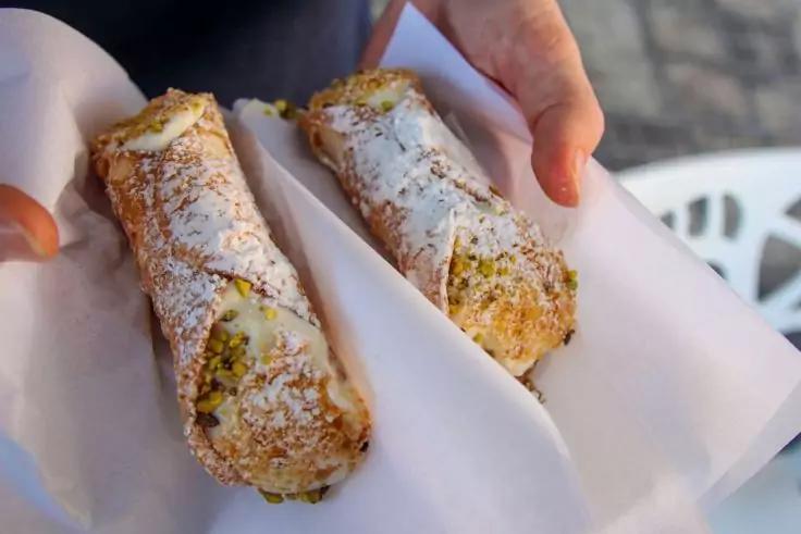 Zwei mit Puderzucker best&auml;ubte Cannoli mit heller F&uuml;llung und gehackten Pistazien an den Enden, auf wei&szlig;en Servietten von H&auml;nden gehalten