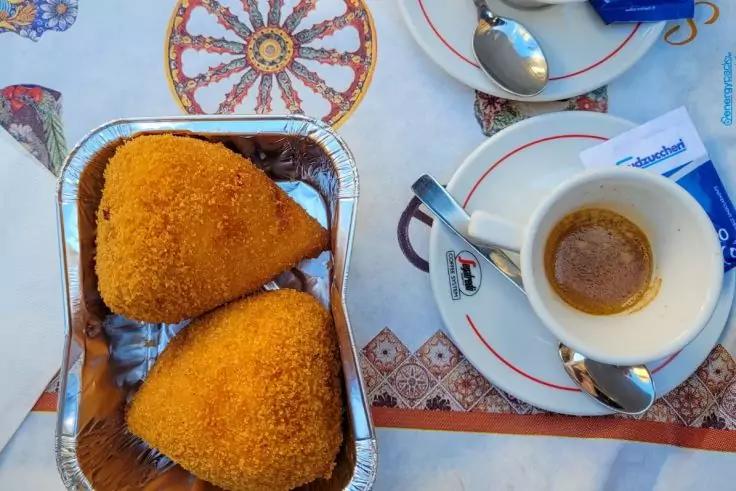 Zwei goldbraune, dreieckige, panierte St&uuml;cke in einer Aluminiumschale; daneben eine kleine Tasse mit dunkler Fl&uuml;ssigkeit auf einer Untertasse, ein L&ouml;ffel und Zuckerp&auml;ckchen auf einer gemusterten Tischdecke