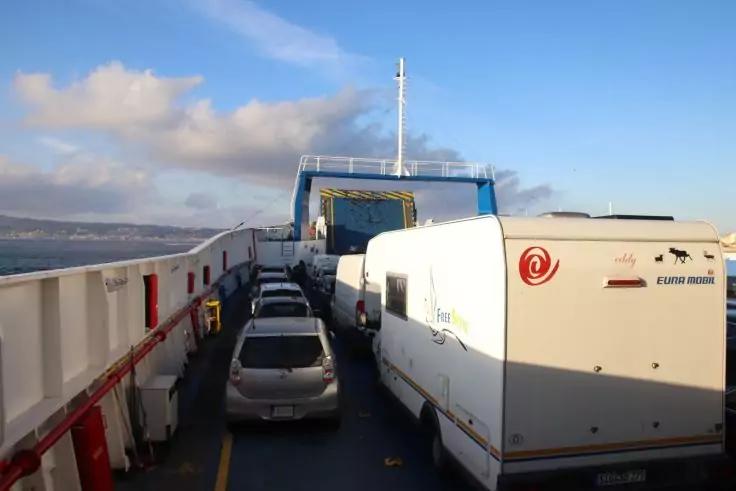 Autos und Wohnwagen auf dem Fahrzeugdeck einer F&auml;hre, Blick nach vorn auf die Schiffsaufbauten, K&uuml;ste links, blauer Himmel mit Wolken