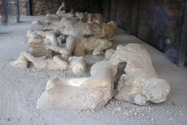 Mehrere wei&szlig;e Gipsabg&uuml;sse in Menschenform liegen auf staubigem Boden unter einem Unterstand; Steinwand im Hintergrund