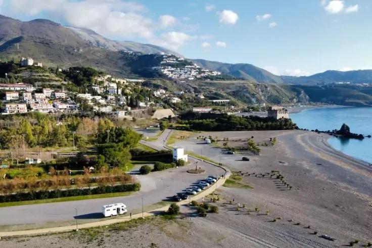 Kiesstrand mit K&uuml;stenstra&szlig;e, geparktem Wohnmobil und Autos, H&auml;usern am Hang, Bergen und wolkigem Himmel