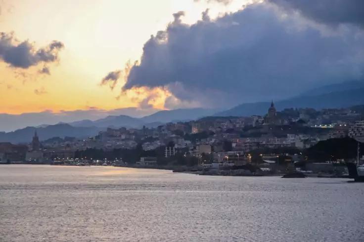 Wasser im Vordergrund, Ufer mit Stadtgeb&auml;uden und vereinzelten Lichtern, H&uuml;gel und Berge im Hintergrund, wolkiger Himmel mit orangefarbenem Licht am Horizont