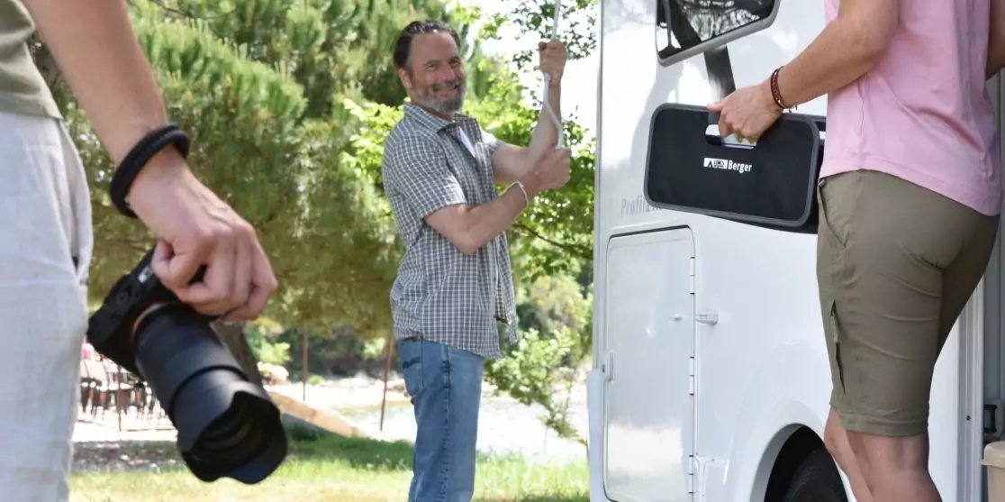 Drei Personen neben einem wei&szlig;en Lieferwagen im Freien. Links im Vordergrund h&auml;lt eine Person eine Kamera, mittig steht eine Person an der ge&ouml;ffneten Seitenklappe des Lieferwagens und ber&uuml;hrt die Kante, rechts h&auml;lt eine Person eine schwarze Tasche