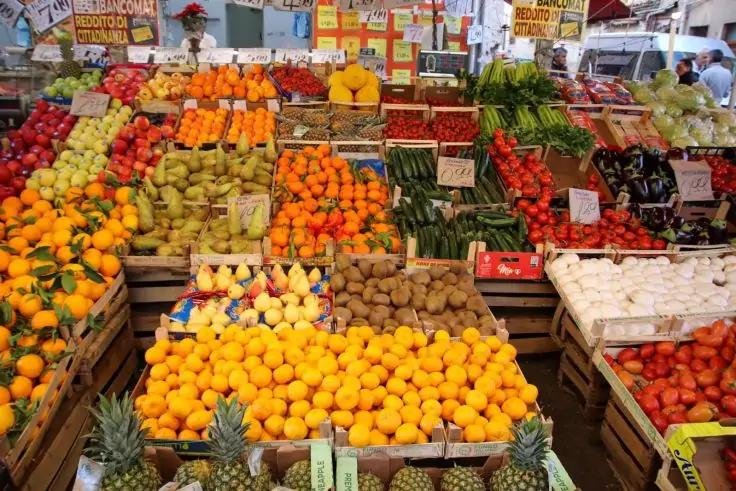 Marktstand mit in Holzkisten aufgereihter Fr&uuml;chte und Gem&uuml;se: Ananas, gelbe Zitrusfr&uuml;chte, Orangen, gr&uuml;ne Birnen, rote &Auml;pfel, Gurken und Tomaten; Preisschilder im Hintergrund
