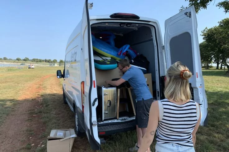 Wei&szlig;er Transporter mit offenen Heckt&uuml;ren auf einer Wiese; zwei Personen am Laderaum; innen bunte aufblasbare Ringe, Kartons, ein Metallbeh&auml;lter und weitere Ausr&uuml;stungsgegenst&auml;nde sichtbar