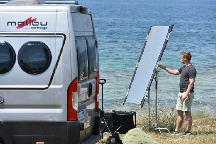 Wei&szlig;er Camper-Van mit zwei runden Fenstern links, gro&szlig;es Solarpanel auf St&auml;nder, ein Mann in dunklem T-Shirt und hellen Shorts ber&uuml;hrt das Panel, grasige K&uuml;stenlinie und Meer im Hintergrund