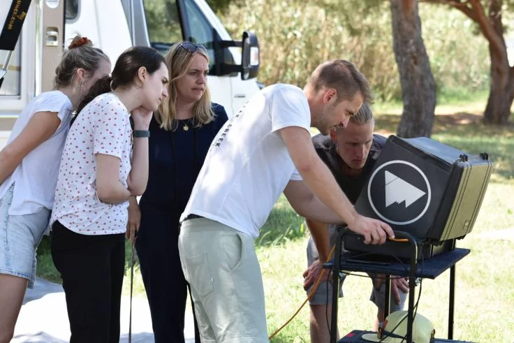 F&uuml;nf Personen stehen im Freien neben einem wei&szlig;en Transporter; eine Person bedient einen offenen Ger&auml;tekoffer mit Bildschirm auf einem Stativ, die anderen sehen zu; B&auml;ume im Hintergrund