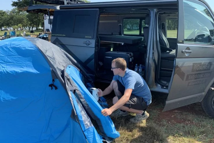 Person mit blauem T‑Shirt und Sonnenbrille kniet neben einem blauen Zelt und h&auml;lt dessen &Ouml;ffnung; daneben ein grauer Van mit ge&ouml;ffneter Schiebet&uuml;r, Grasfl&auml;che und B&auml;ume im Hintergrund
