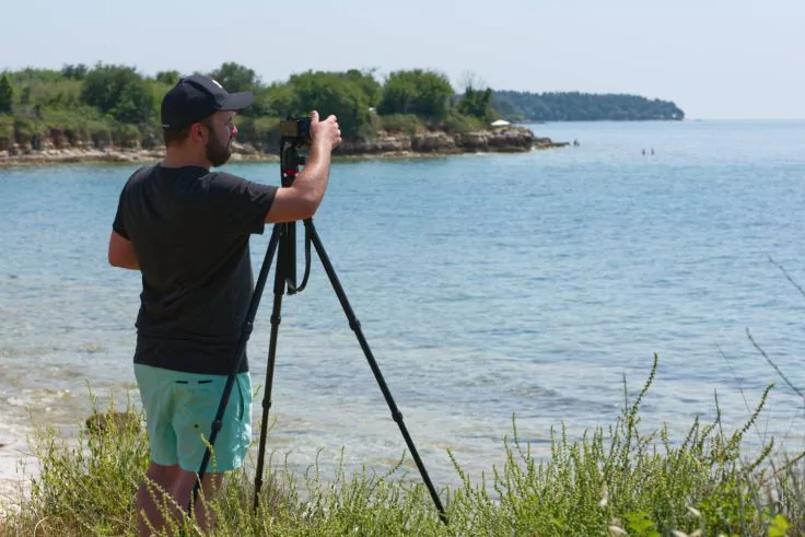Person mit Kappe bedient eine Kamera auf einem Stativ am grasigen Ufer; im Hintergrund Felsen, K&uuml;stenvegetation und ruhiges Meer, zwei Personen im Wasser