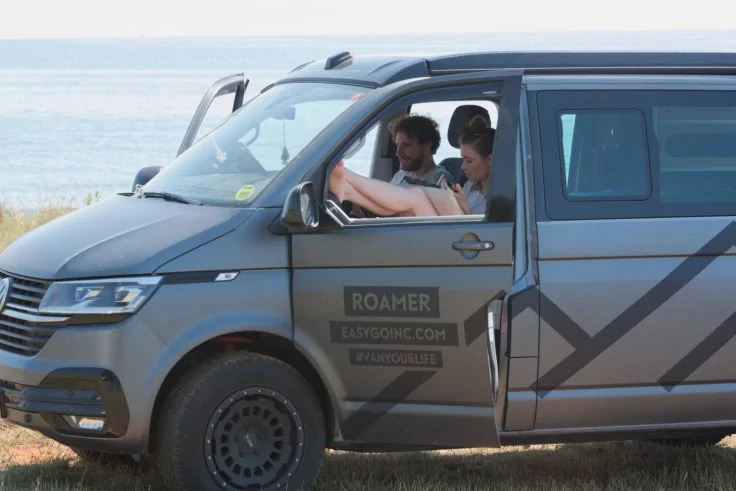 Grauer Camping-Van mit ge&ouml;ffneter Schiebet&uuml;r, zwei Personen vorne schauen auf ein Buch, geparkt auf Gras am Meer; Schriftzug 'ROAMER' auf der T&uuml;r