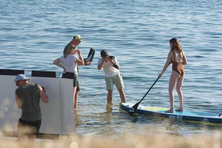 Person im Bikini auf einem Stand-up-Paddle-Board im flachen Wasser; mehrere Personen mit Kameras und Reflektor daneben; Sandstrand im Vordergrund, Meer im Hintergrund